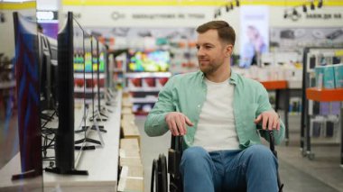 Tekerlekli sandalye kullanıcısı bir elektronik mağazasında televizyon modellerini karşılaştırıyor, elinde bir kağıt parçası, muhtemelen bir alışveriş listesi ya da ürün özellikleri tutuyor.