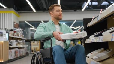 Profesyonel tekerlekli sandalye kullanıcısı elektronik cihazlara göz atıyor, gülümserken özgüvenle projektör seçiyor, modern perakende ortamda bağımsız yaşam ve ulaşılabilirliği temsil ediyor