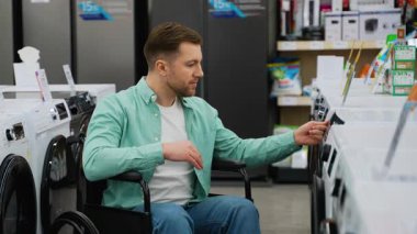 Tekerlekli sandalye kullanıcısı elektronik mağazasında geziniyor, bağımsız olarak çamaşır makinesi özelliklerini karşılaştırıyor ve özerklikle düşünceli satın alma kararı veriyor