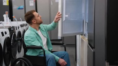Tekerlekli sandalyedeki adam bir ev aletleri mağazasında çeşitli buzdolabı ve diğer aletleri keşfediyor, engelli insanlar için alışveriş yapmanın ulaşılabilirliğini gösteriyor.