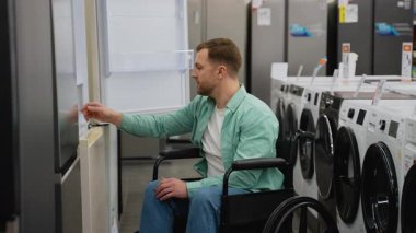 Tekerlekli sandalyedeki adam yeni buzdolabının özelliklerini keşfediyor, ev aletleri mağazasındaki aletleri inceliyor, bağımsız yaşam ve tüketici seçimini gösteriyor