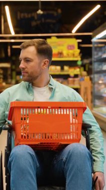 Süpermarkette maluliyet alışverişi yapan mutlu bir adam, tekerlekli sandalyede oturuyor ve raflara bakıyor, bacaklarında alışveriş sepeti tutuyor, koridorda yürüyor.