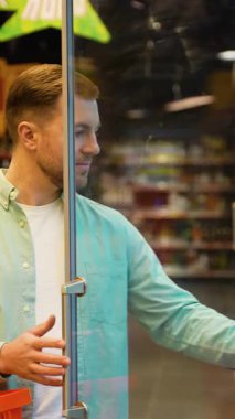 Marketin girişine odaklanmış müşteri, alışveriş sepetini kavrıyor ve cam kapıdan perakende piyasaya geçerken su şişesini dikkatle inceliyor.