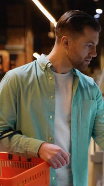 Turuncu alışveriş sepeti tutarken vakumlu paketlenmiş somon etiketini dikkatlice inceleyen genç adam, sağlıklı protein seçenekleri için marketlerin soğutulmuş kısımlarına bakıyor.