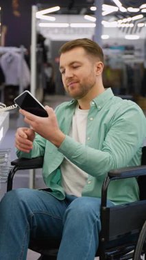 Tekerlekli sandalyede oturan mutlu adam, elektronik mağazasında yeni akıllı telefonu test ediyor, cep telefonu seçiyor ve satın alıyor, engelliler için ulaşılabilir bir ortam.