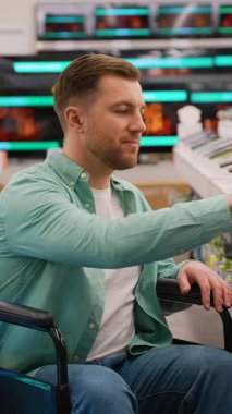 Tekerlekli sandalye kullanıcısı elektronik mağazasında geziniyor, akıllı telefon kullanarak televizyon modellerini ve fiyatlarını karşılaştırırken aynı zamanda teknoloji erişilebilirliğini ve bağımsız tüketici seçimini gösteriyor