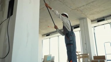 Üniforma giyen profesyonel bir ressam, paten fırçası kullanarak duvara boya sürüyor ve kapalı bir inşaat alanında özenle çalışıyor.