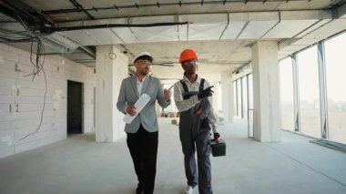 İnşaat işçisi ve mimar işbirliği yapıyor, kısmen tamamlanmış binada yürüyor, mimari planları inceliyor ve proje geliştirmeyi tartışıyor