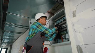 Profesyonel teknisyen elektrik paneli bakımı yapıyor, bağlantıları dikkatlice kontrol ediyor ve özel araçlar ve koruyucu ekipmanlarla endüstriyel çalışma alanındaki güvenlik protokollerini uyguluyor.
