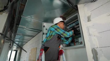 Güvenlik teçhizatı takan profesyonel elektrikçi modern inşaat alanındaki elektrik panelini dikkatlice döşeyerek elektrik sistemi bütünlüğünü ve işçi güvenliğini koruyor.