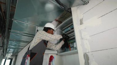 Üniformalı profesyonel elektrikçi, ticari binalardaki kabloları dikkatle denetliyor ve ayarlıyor. Elektrik paneli, güvenlik ve işlevselliği sağlıyor.