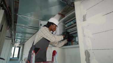 Koruyucu teçhizat kullanan elektrikçi endüstriyel bir ortamda devre kesici panelinde çalışıyor. Titiz kurulum ve bakım tekniklerini gösteriyor.