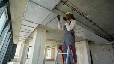 Profesyonel elektrikçi merdiven üzerinde çalışıyor. İnşaat halindeki bir binanın tavanına dikkatlice elektrik kabloları yerleştiriyor. Güvenliği ve yönetmeliklere uymayı sağlıyor.