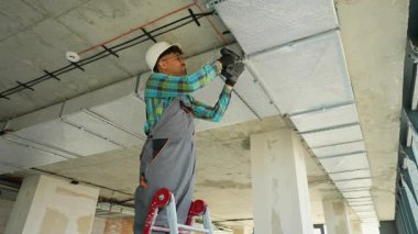 Güvenlik kaskı ve üniforması giyen profesyonel elektrikçi merdivenin üzerinde duruyor ve inşaat halindeki bir binanın tavanına elektrik kabloları yerleştiriyor.