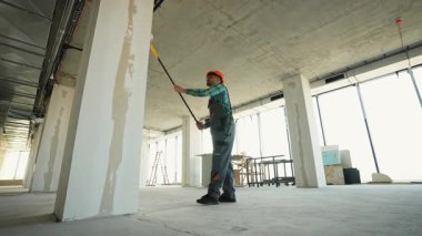 Üniformalı ve şapkalı inşaat işçisi inşaat halindeki modern ofis binasının duvarlarına astar astar takarak son boyaya hazırlanıyor.