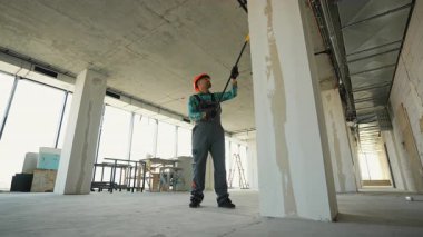 Profesyonel inşaat işçisi iç duvarları ve sütunları uzun paten fırçasıyla dikkatlice boyuyor, bina yapısını yenilerken koruyucu kask ve üniforma giyiyor.