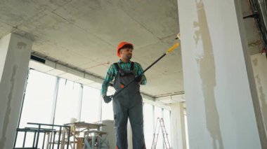 Profesyonel inşaatçı inşaat halindeki bir binanın duvarlarını ve sütunlarını uzun bir fırça kullanarak boyamaya hazırlanıyor.