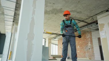 Profesyonel müteahhit, tadilat altındaki bir dairede duvarları ve tavanları sıva ile kaplıyor, uzun bir spatula kullanıyor ve koruyucu iş kıyafeti giyiyor.