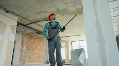 Üniforma ve şapka giyen profesyonel müteahhit, duvarlara bir silindir kullanarak astar takıyor ve onları yeni bir dairenin yenilenmesi sırasında boyamaya hazırlıyor.