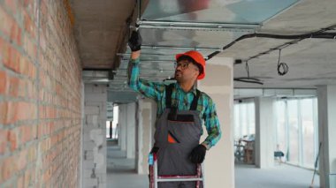 Güvenlik teçhizatı takan yetenekli elektrikçiler bitmemiş binanın tavanına kablo döşüyorlar, profesyonel elektrik altyapı çalışmalarını vurguluyorlar.