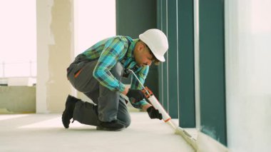 Profesyonel müteahhit, inşaat halindeki modern bir binada hava geçirmez ve su geçirmez bir tesis sağlamak için dolgu tabancası kullanarak geniş bir pencere çerçevesini mühürlüyor.