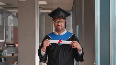 Mezuniyet elbisesi ve şapkası giymiş gururlu bir öğrenci, diplomasını tatmin olmuş bir gülümsemeyle gösteriyor, akademik başarısını kutluyor ve çalışmalarını başarıyla tamamlıyor.