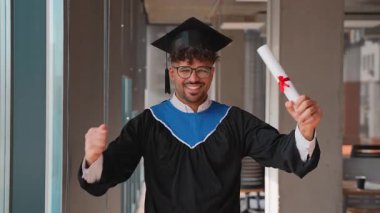 Siyahi mezuniyet elbisesi ve kepiyle üniversite koridorunda dikilmiş gururla diplomasını tutuyor ve akademik başarısını neşeli bir ifadeyle kutluyordu.