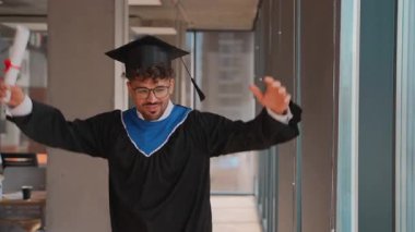 Mezuniyet elbisesi ve şapkası giymiş genç mezun, elinde diplomasıyla bir üniversite koridorunda neşe ve başarısını ifade ediyor.