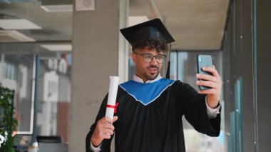 Eğitim kilometre taşını kutlayan resmi akademik giysiler içinde gururlu bir mezun, diploma sergilerken akıllı telefon kullanan sevdikleriyle video görüşmeleri yoluyla başarıyı paylaşıyor