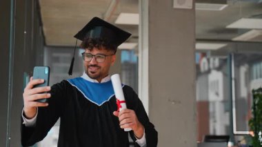 Akademik regalia giyip mezun olmuş, başarıyı kutluyor, diplomasını tutuyor ve kampüs ortamından akıllı telefondan telefon görüşmesi yapıyor.