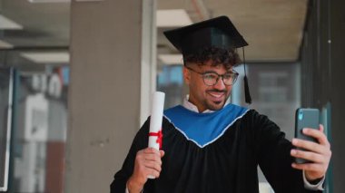 Mezuniyet kıyafetiyle mezun olan genç latin, elinde gururla diplomayla akıllı telefondan görüntülü konuşma yapıyor, akademik başarıyı ve gelecek kariyer beklentilerini kutluyor.