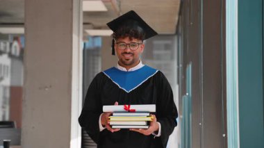 Genç mezun koridorda duruyor, tören elbisesi ve mezuniyet şapkası takıyor, diploma ve ders kitaplarını tutuyor, akademik başarılarını samimi bir gülümsemeyle ifade ediyor.