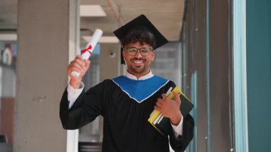 Diploma ve ders kitaplarını gururla elinde tutarken, kampüs koridorunda neşe ve akademik başarı saçan kendinden emin üniversite mezunu.