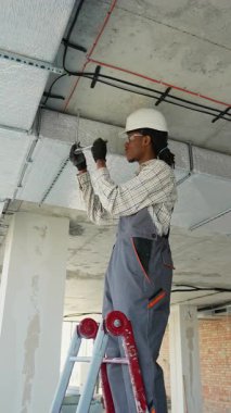 Güvenlik gözlüğü ve miğfer takan profesyonel elektrikçi, merdivenin üzerinde duruyor, inşaat halindeki bir binanın tavanına dikkatlice elektrik kabloları yerleştiriyor.