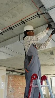 Güvenlik gözlüğü ve miğfer takan profesyonel elektrikçi, merdivenin üzerinde duruyor, inşaat halindeki bir binanın tavanına dikkatlice elektrik kabloları bağlıyor.