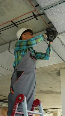 Merdivende çalışan profesyonel elektrikçi inşaat halindeki bir binanın tavanına elektrik kanalı kurmak için matkap kullanıyor.