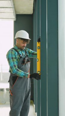 Korumalı güvenlik teçhizatı takan inşaat işçisi, yeşil duvar yüzeyini dikkatlice ölçüyor, sarı ruh seviyesini kullanarak, bina bakımında profesyonel hassasiyeti gösteriyor.