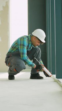 Profesyonel müteahhit, zemin eklemlerini dolgu tabancasıyla dikkatlice mühürlüyor, güvenlik kaskı takıyor ve iş tulumu giyiyor, parlak iç dekorasyon düzeninde kusursuz inşaat becerisi gösteriyor.