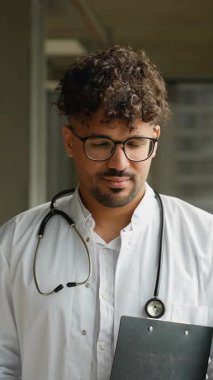 Kıvırcık saçlı, gözlüklü ve boynunda steteskop olan genç bir erkek doktor bir pano tutuyor ve düşünceli bir gülümsemeden geniş bir gülümsemeye kadar birçok yüz ifadesi sergiliyor.