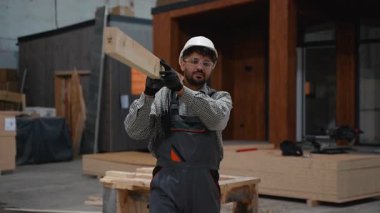 Usta inşaat işçisi atölye boyunca ahşap kalas taşıyor, koruyucu giysiler giyiyor ve profesyonel marangozluk yapıyor.