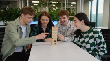 Dört beyaz öğrenci, iki erkek ve iki kadın, beyaz bir masanın etrafında toplanmış, Jenga oyununa dalmış, dikkatle ahşap blokları teker teker kaldırıyorlar.