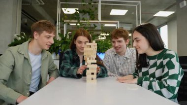 Kafkas üniversite öğrencileri stratejik olarak Jenga oynuyorlar, tahta blokları beyaz masa etrafında odaklanmış heyecan ve rekabetçi ruh ile dengelemekteler.