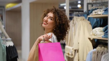 Giyim mağazasında duran genç bir kadın, elinde geniş bir gülümsemeyle pembe bir alışveriş çantası tutuyor, başarılı alışveriş deneyiminden ve yeni moda alışverişinden duyduğu memnuniyeti dile getiriyor.