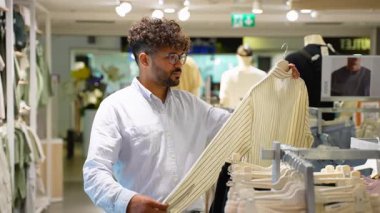 Odaklanmış genç erkek alışverişçi elbise raflarını karıştırıyor, perakende mağazasından alışveriş yaparken gömlek kumaşını, formasını ve kalitesini titizlikle inceliyor.
