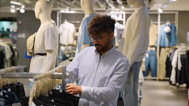 Giyim mağazasında kot pantolon seçen genç erkek alışverişçi, farklı giyim seçeneklerini karşılaştırırken beden, renk ve uygun mu diye dikkatlice kotları inceliyor.