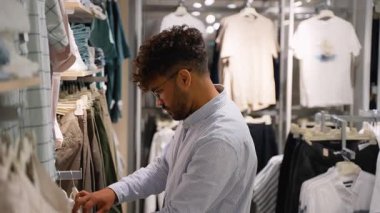 Modern bir giyim mağazasında çeşitli kıyafetleri dikkatlice inceleyen genç bir erkek müşteri, satın almadan önce farklı stil ve renkleri göz önünde bulundurarak