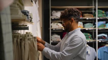 Genç erkek alışverişçi giyim mağazasında pantolon seçerken, farklı stil ve renkleri karşılaştırırken erkek giyim koleksiyonunu dikkatle inceliyor.