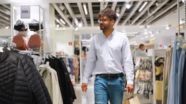 Genç sakallı erkek alışverişçi giyim mağazasına göz atıyor, alışveriş torbası taşırken perakende satış raflarından şık ürünler seçiyor.