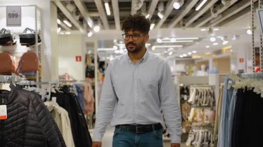 Kendine güvenen genç ortadoğulu bir adam giyim mağazasından geçiyor raflardaki çeşitli kıyafetlere bakıyor, gülümsüyor ve alışveriş deneyiminin tadını çıkarıyor.