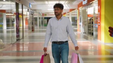 Mutlu Arap adam gözlük takıyor ve gündelik giysiler giyiyor. Parlak bir alışveriş merkezinde elinde poşetlerle dolaşıyor. Başarılı alışveriş çılgınlığının tadını çıkarıyor.